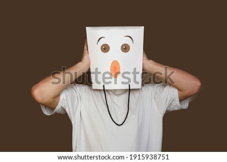 Young man hiding face behind paper bag with drawn emoticon on light background