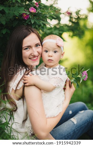 Mother and child daughter in spring garden. Young woman and baby girl together standing near the blooming bush with pink rose flowers. Baby touching flowers on bush. summer time.