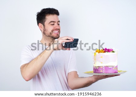 Young handsome Caucasian smiling man standing against white background taking a picture with his smartphone to a beautiful pink flowered designed cake and posting it to his social media accounts.