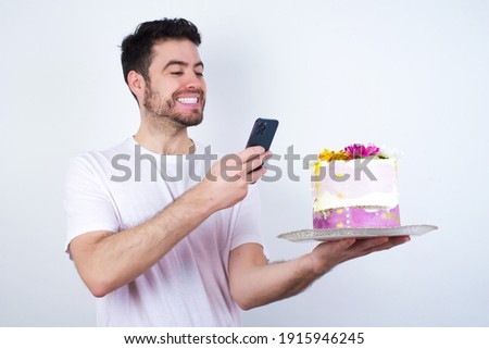 Young handsome Caucasian smiling man standing against white background taking a picture with his smartphone to a beautiful pink flowered designed cake and uploading it on his social media