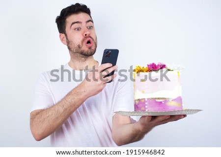 Young handsome Caucasian surprised man standing against white background taking a picture of a beautiful pink flowered designed cake ready to post it on social media or send it to his friends. 