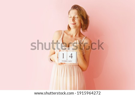 Pretty woman is holding date calendar with 14 February Happy in front of pink background Valentine Day