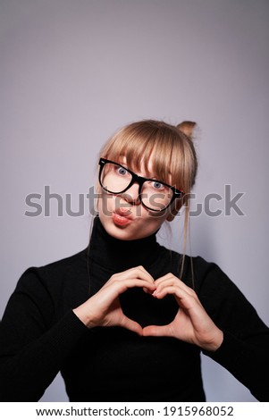 Young blonde woman wearing black sweater and glasses showing heart symbol and shape with hands isolated on gray background. Romantic concept