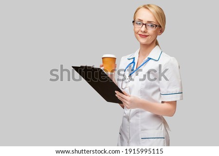 Portrait of a female doctor holding paper cup of coffee and holding tablet pc pad standing over grey background