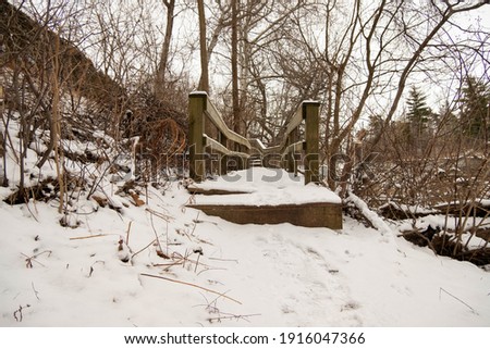 Snow covered wooden path in the forest in London Ontario, Canada. Cold winter day with gloomy sky and bare brown trees.