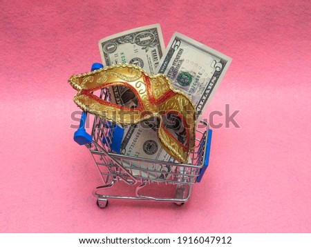 A traditional Venetian golden carnival mask with money (dollar cuppures) in a toy shopping cart on a pink background. Concept of preparation for the annual Brazilian festival, costume rental.