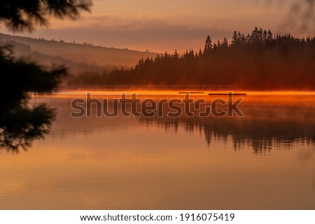 Magical morning in nature in the reddish light of sunrise on a quiet mountain lake surrounded by coniferous forest and covered by fog floating calmly above the water