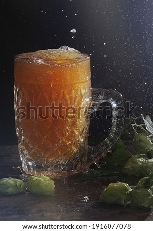 Splashes and puddles next to a large mug with poured beer on a black background with sprouts of hops.