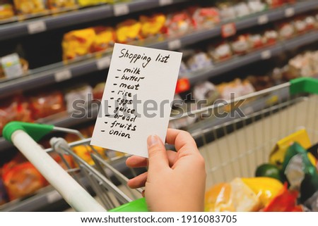 Woman with notebook in grocery store, closeup. Shopping list on paper. Check purchases in grocery cart. Royalty-Free Stock Photo #1916083705