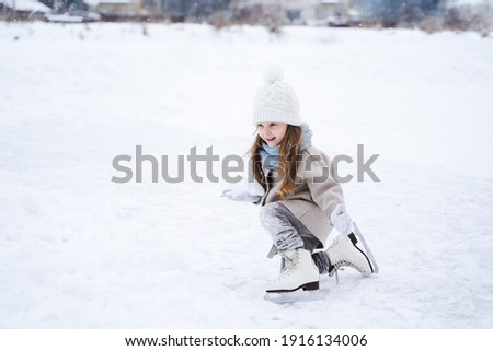 Beautiful little cute girl learn to skate on ice skating rink in park. Fall down and have fun. Stylish look, warm woolen coat, white hat, scarf, snood. Winter family activities, sport, games outdoors.