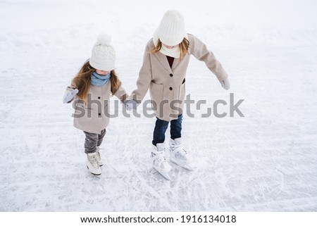 Beautiful little girls, sisters learn to skate on ice skating rink in park. Fall and have fun. Stylish looks, warm woolen coats, white hats, scarves, snoods. Winter family activities, games outdoors.