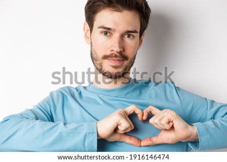Close-up of young romantic man with beard, showing heart gesture and looking at camera, show I love you sign, white background