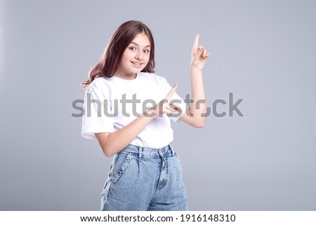 Cute girl makes a pointing gesture with her hands. Emotional face. Advertising, great idea concept. Dressed in white T-shirt, jeans. Studio light. Grey background. Caucasian. Copy space.