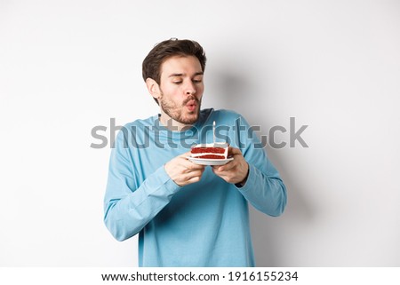 Celebration and holidays concept. Happy young man blowing candle on birthday cake, making wish on bday, standing over white background