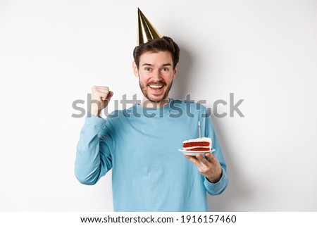 Celebration and holidays concept. Cheerful young man celebrating birthday in party hat, holding bday cake and looking happy, standing on white background