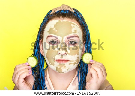 Woman with blue afro braids face in a mask made of green clay with cucumber circles in her hands on a yellow background. Horizontal photo