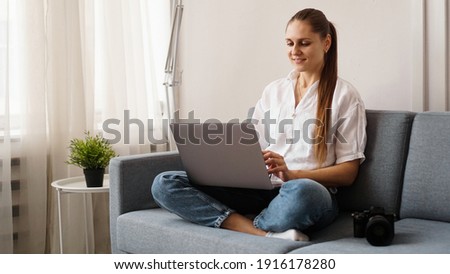 Happy young woman using laptop at home. On the couch next to the woman is a camera. Photographer retouches photographs at home.
