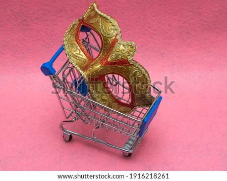 Traditional gold Venetian carnival mask toy mini shopping cart (trolley) on a pink background. The concept of preparing for the annual Brazilian festival, buying costumes.