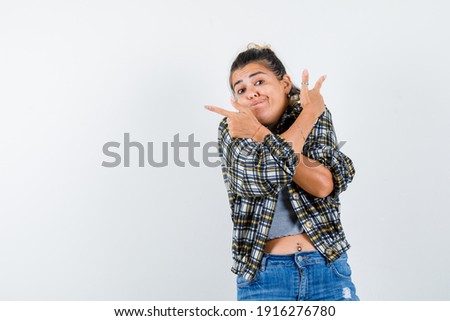  young lady keeping crossed arms on chest while pointing up in shirt,jeans and looking hesitant , front view. 