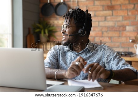 Close up smiling dreamy African American man wearing headphones sitting at table with laptop, looking to aside, dreaming about new opportunities, satisfied businessman working online at home Royalty-Free Stock Photo #1916347676