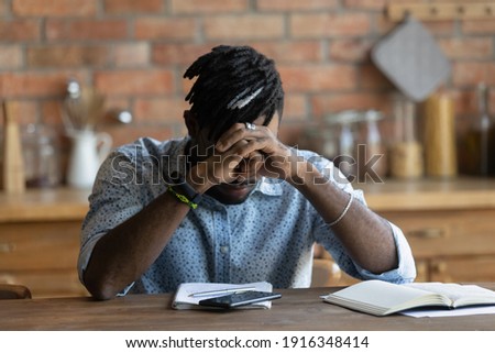 Close up depressed exhausted African American man sitting at table with head down, received message with bad news, thinking about problems, bankruptcy or dismiss, lost job, feeling nervous Royalty-Free Stock Photo #1916348414