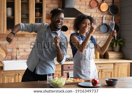 Close up overjoyed African American couple singing into kitchenware, having fun, dancing, listening to music in kitchen, cooking salad, excited wife and husband holding whisk and ladle a microphone Royalty-Free Stock Photo #1916349737