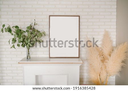 Stylish elegant eco composition of living room with white false fireplace, empty picture frame, dried flowers plants pampas grass. Modern home decor. Template. Mock up poster frame on the wall.