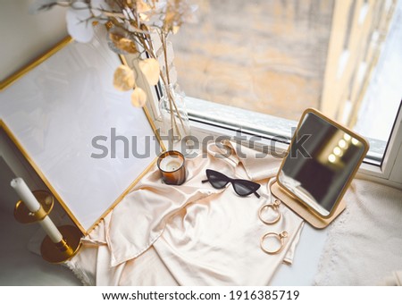 Cozy spring still life feminine scene golden shades. Female styled window sill minimalistic composition. Empty picture mock up poster frame, elegant accessories mirror earrings, dried flowers, candle