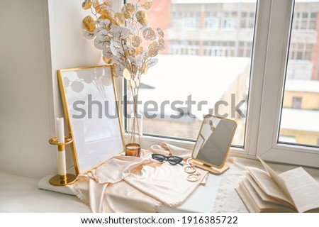 Cozy spring still life feminine scene golden shades. Female styled window sill minimalistic composition. Empty picture mock up poster frame, elegant accessories mirror earrings, dried flowers, candle