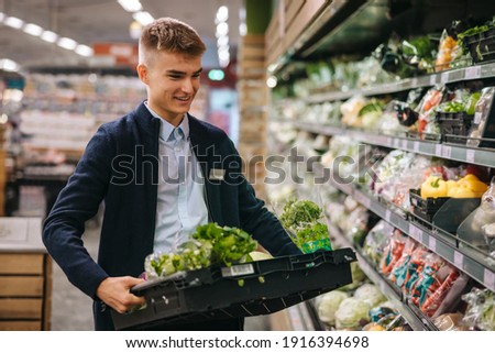 Shop assistant in supermarket re-stocking fresh vegetables in shelves of produce section.