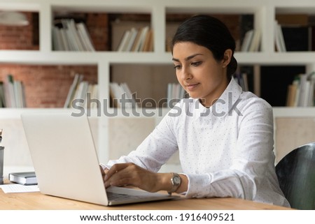 Concentrated young Indian female worker sit at desk look at laptop screen consult client or customer online. Mixed race ethnicity woman employee type text on computer in office. Technology concept. Royalty-Free Stock Photo #1916409521