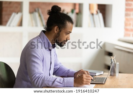 African American male employee sit at desk in home office work on laptop online with paper documents. Pensive mixed race ethnicity man study on computer, take notes. Distant education concept.