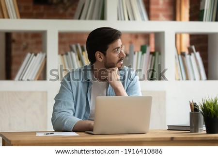 Pensive young Caucasian businessman sit at desk in office work on laptop look in distance solving business problem. Thoughtful male employee distracted from computer job think or plan at workplace. Royalty-Free Stock Photo #1916410886