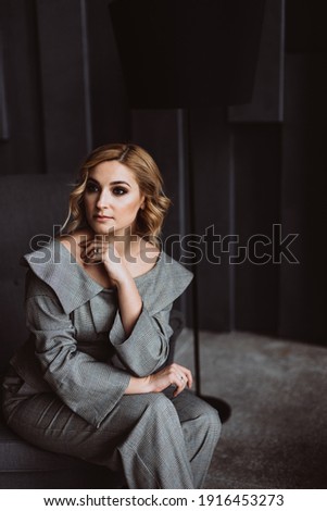 Beautiful young businesswoman in a stylish gray suit sitting on a chair in an office with a modern interior, looking at the camera. Portrait of a charming business woman. Soft selective focus.