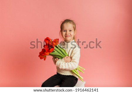 happy little blonde girl sitting and holding red tulips on pink background with space for text