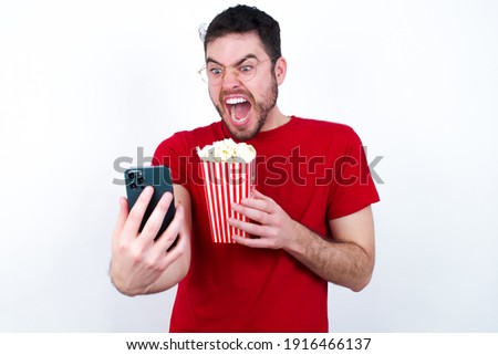 Photo of outraged annoyed Young handsome man in red T-shirt against white background eating popcorn holds cell phone, makes call, argues with colleague,  expresses negative emotions. People and anger.