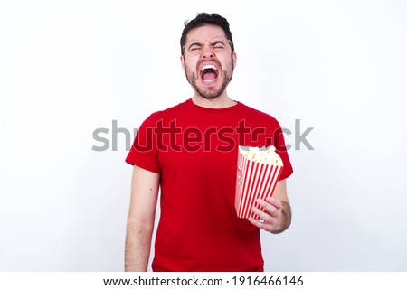 Stressful Young handsome man in red T-shirt against white background eating popcorn screams in panic, closes eyes in terror, keeps hands on head, finds out terrified news, can't believe it.
