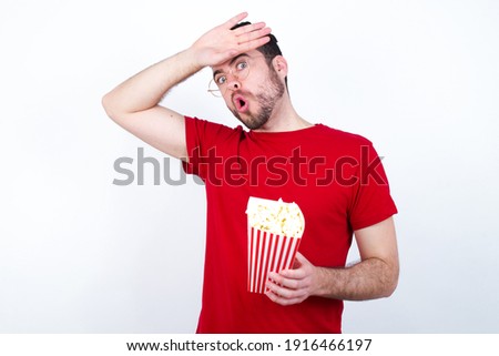 Young handsome man in red T-shirt against white background eating popcorn wiping forehead with hand making phew gesture, expressing relief feels happy that he prevented huge disaster. 