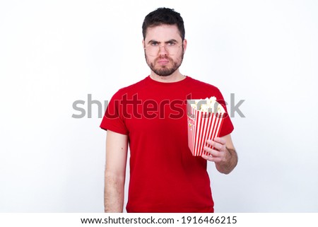 Young handsome man in red T-shirt against white background eating popcorn frowning his eyebrows being displeased with something.
