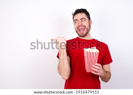Stupefied Young handsome man in red T-shirt against white background eating popcorn with surprised expression, opens eyes and mouth widely, points aside with thumb, shows something strange.