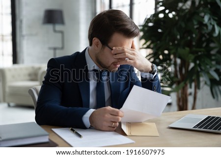 Upset young Caucasian businessman feel depressed frustrated by bad news in paper document. Unhappy sad male CEO distressed by negative response or message in paperwork letter or correspondence. Royalty-Free Stock Photo #1916507975