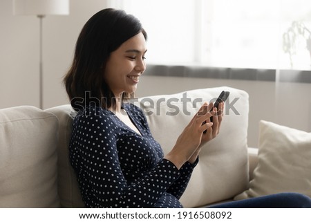 Smiling vietnamese female spend leisure time on cozy couch at home browsing social networks online reading blogs news using cell. Tranquil young asian woman watching video photo on smartphone screen