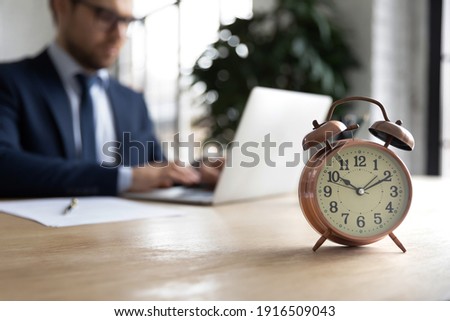 Close up of clock on forefront with businessman work on laptop online in office on background. Male employee prepare project at workplace, try meet deadline or appointment. Time management concept. Royalty-Free Stock Photo #1916509043