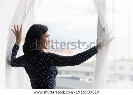 Happy excited young asian female tourist traveler opening curtains on large window of luxury hotel suite enjoying urban view. Amazed millennial vietnamese woman meeting morning at new flat apartment