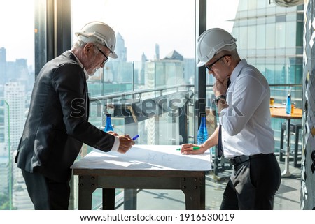 Two engineers discuss their construction plan for their new site. They had a drawing board with pen in their hand. They both wear hard hat. They are working near the building they are constructing.