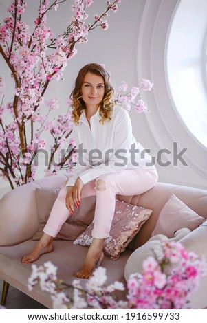 Portrait of a beautiful young girl with blond hair on a background of pink sakura flowers. She sits on the back of the sofa. Selective focus of the image.