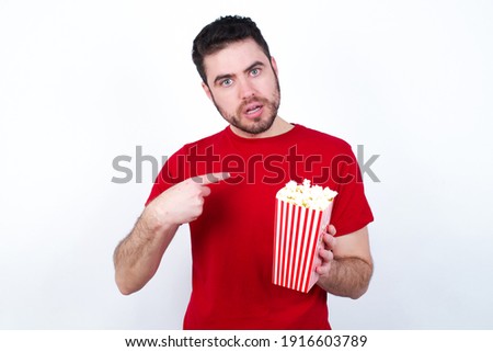 Embarrassed Young handsome man in red T-shirt against white background eating popcorn indicates at herself with puzzled expression, being shocked to be chosen to participate in competition, hesitates 
