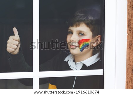 Lonely school kid boy sitting by window with rainbow with colorful colors on face. Child during pandemic coronavirus quarantine. Children make and paint rainbows around the world.
