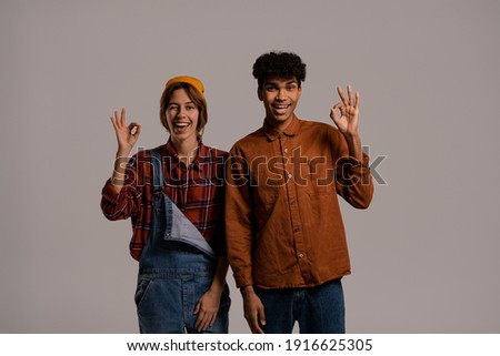 Photo of cute couple farmers show OK and look happy together. White woman wears denim overall, black man shirt isolated grey color background