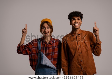 Photo of cute couple farmers smile and point up. White woman wears denim overall, black man shirt isolated grey color background